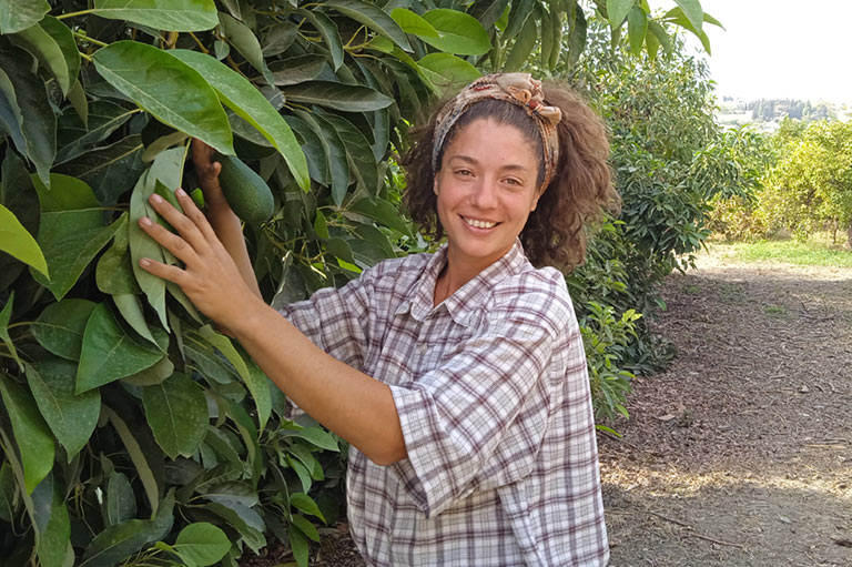 Belén, staand naast een avocadoboom, met een van de groeiende avocado's in de hand